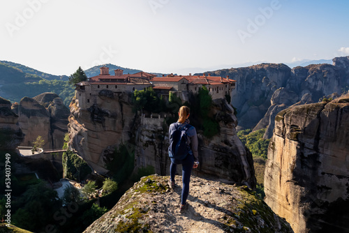 Tourist woman standing on cliff edge with scenic view of Holy Monastery of the Holy Trinity, Kalambaka, Meteora, Thessaly, Greece, Europe. Dramatic landscape. Landmark build on unique rock formations photo