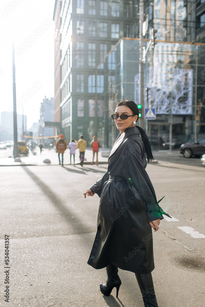 Girl on the street in a black coat. Stylish girl in a black raincoat on the background of a high glass house