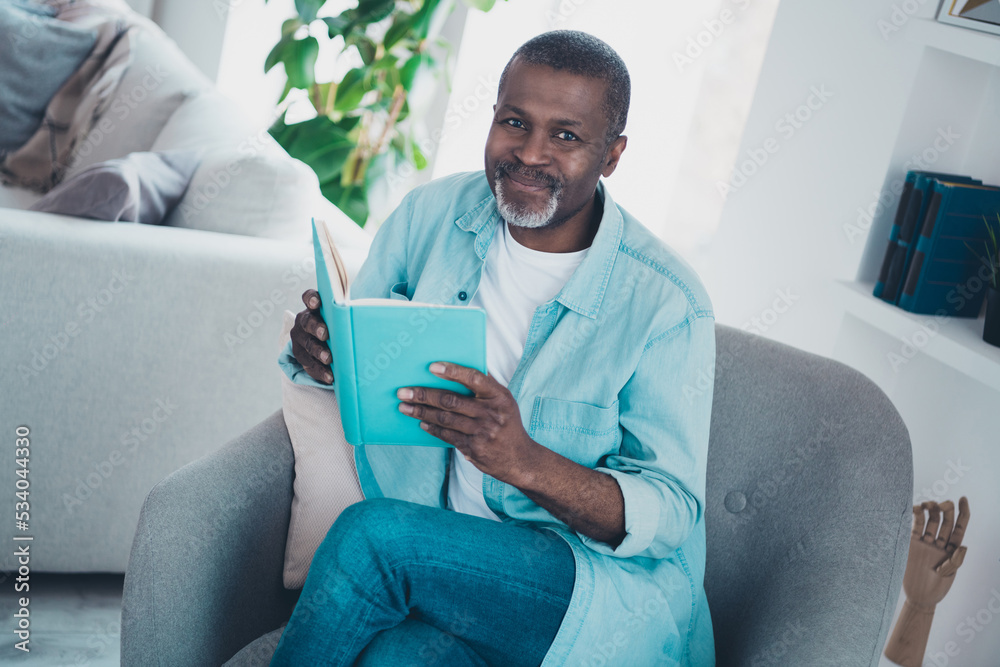 Photo of cheerful positive man sit armchair hold book look camera have good mood pastime house indoors Photo of cheerful positive man sit armchair hold book look camera have good mood pastime house indoors