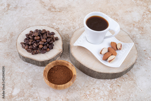 Pile of coffee beans on a wooden board next to a small bowl of ground coffee and a cup of coffee with almonds and pistachios on marble background