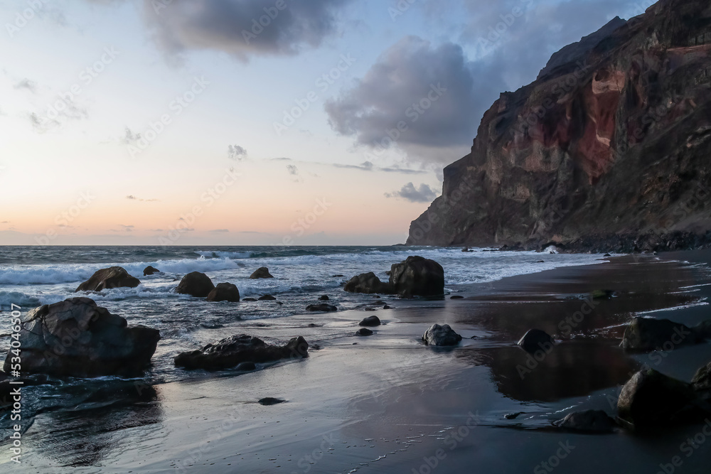 Scenic view during sunset on volcanic black sand beach Playa del Ingles in Valle Gran Rey, La Gomera, Canary Islands, Spain, Europe. Massive cliffs of the La Mercia range. Smooth waves sweeping rocks