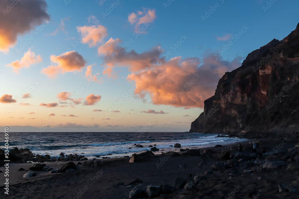 Scenic view during sunset on the volcanic sand beach Playa del Ingles in Valle Gran Rey, La Gomera, Canary Islands, Spain, Europe. Massive cliffs of the La Mercia range. Calm atmosphere at the seaside Scenic view during sunset on the volcanic sand beach Playa del Ingles in Valle Gran Rey, La Gomera, Canary Islands, Spain, Europe. Massive cliffs of the La Mercia range. Calm atmosphere at the seaside