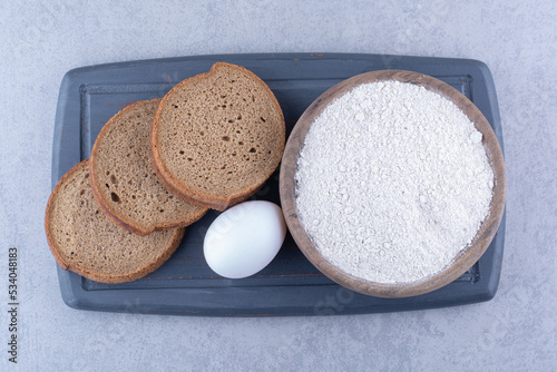 Small bowl of flour, an egg and three bread slices on a navy board on marble background