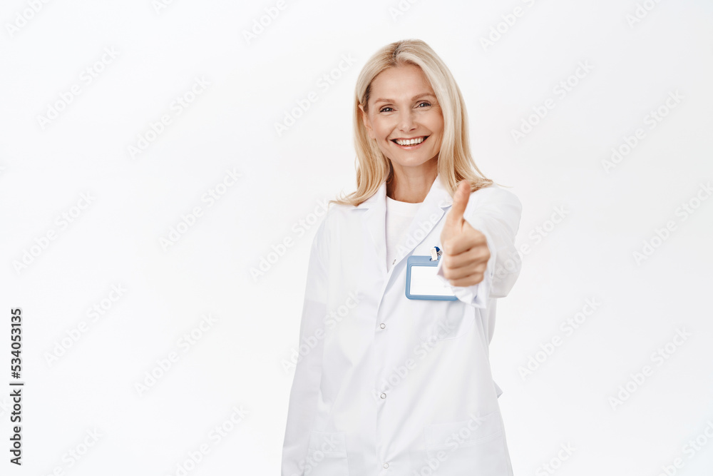 Smiling mature woman doctor, 50s years, shows thumbs up, recommends and promotes healthcare clinic, approves smth, stands over white background Smiling mature woman doctor, 50s years, shows thumbs up, recommends and promotes healthcare clinic, approves smth, stands over white background