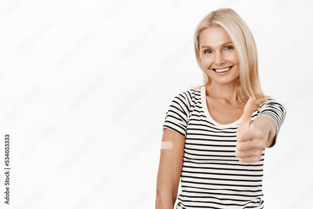 Happy smiling mature woman, 50 years old, has covid-19 vaccination band aid on shoulder, making thumbs up gesture, recommending vaccinate, white background Happy smiling mature woman, 50 years old, has covid-19 vaccination band aid on shoulder, making thumbs up gesture, recommending vaccinate, white background