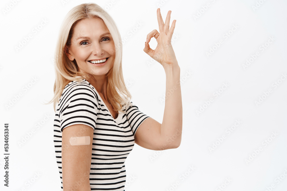 Smiling grandmother did covid-19 vaccination shot, shows her arm with band aid after vaccine, makes okay sign, stands over white background Smiling grandmother did covid-19 vaccination shot, shows her arm with band aid after vaccine, makes okay sign, stands over white background