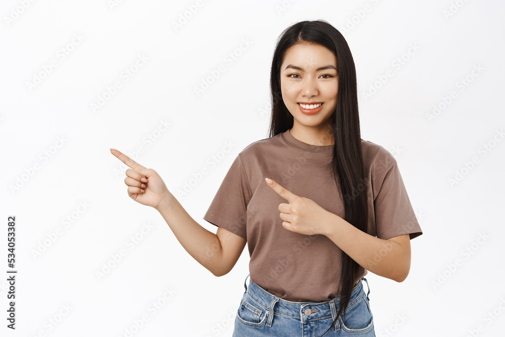 Advertising. Smiling asian woman pointing fingers left, showing promo offer. Chinese girl demonstrates banner, white background Advertising. Smiling asian woman pointing fingers left, showing promo offer. Chinese girl demonstrates banner, white background