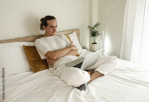Happy young man sitting on bed at home and working on laptop