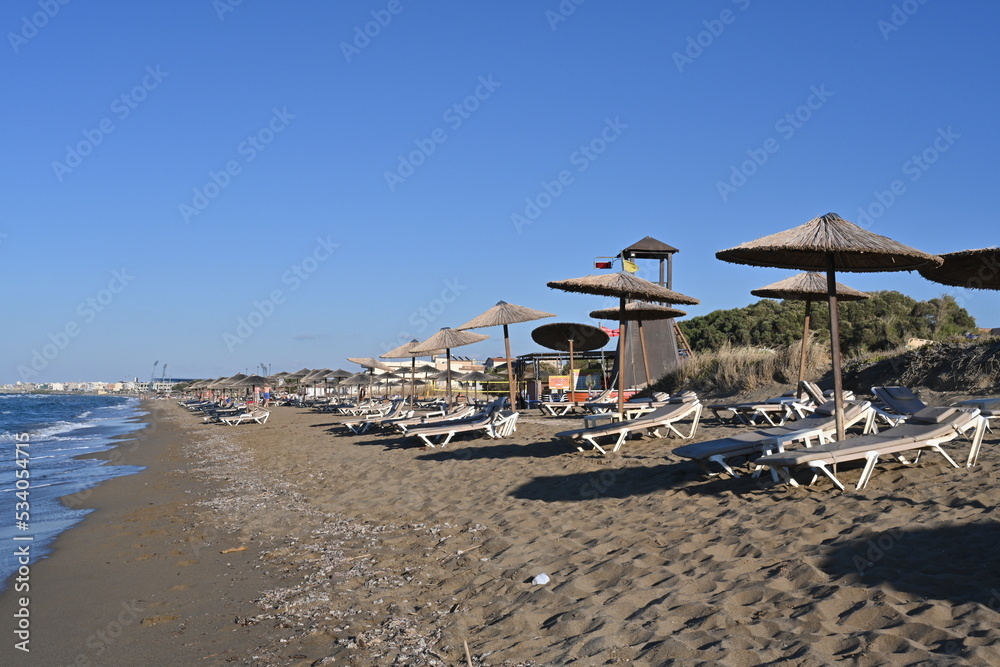 Sandy beach of a holiday resort with plastic deck chairs and straw beach umbrellas. Among them there is a tower for lifeguard. Copy space is available. Cloudless sky is on the background.