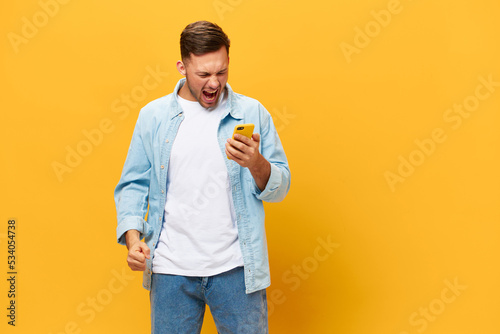 Angry irritated tanned handsome man in blue basic t-shirt scream at smartphone posing isolated on orange yellow studio background. Copy space Banner Mockup. Online Social media concept. Bad news