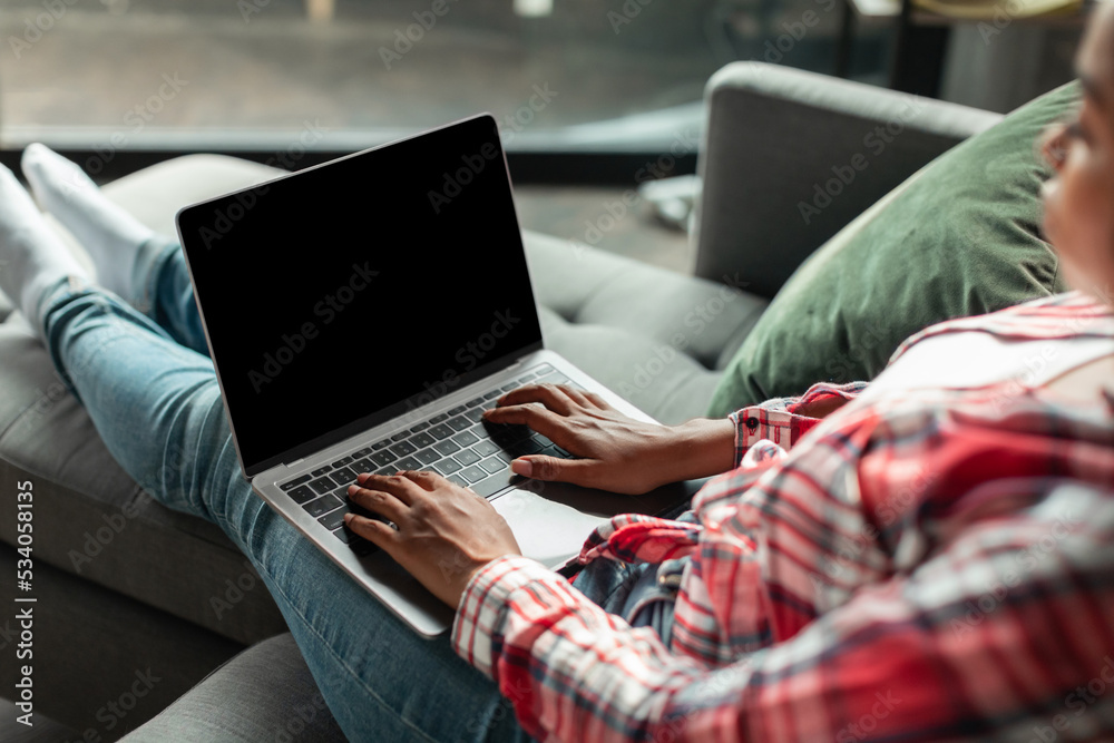 Fototapeta premium Serious millennial african american woman typing on laptop with blank screen on sofa in living room