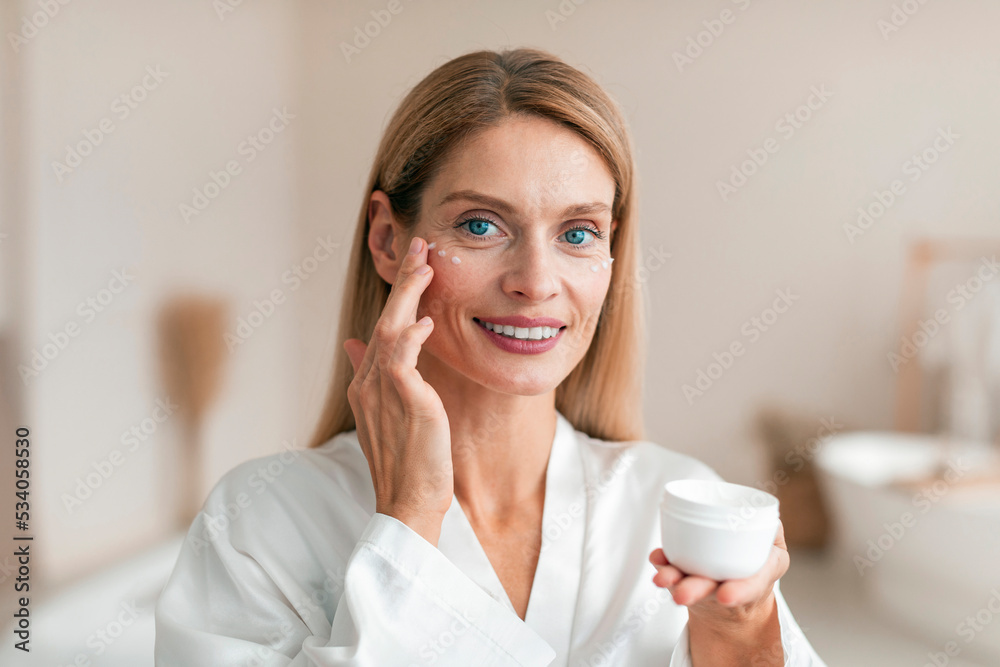 Beautiful middle aged woman in silky bathrobe using moisturizing eye cream, enjoying face care routine - obrazy, fototapety, plakaty 