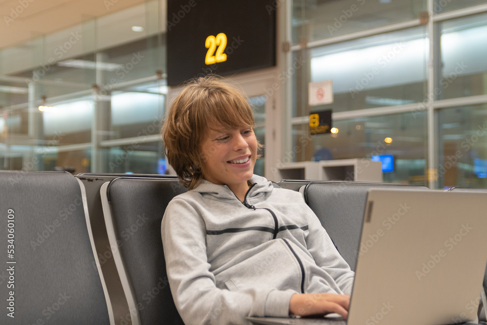 Obraz premium Blond boy waits in an airport lounge while using his laptop computer.