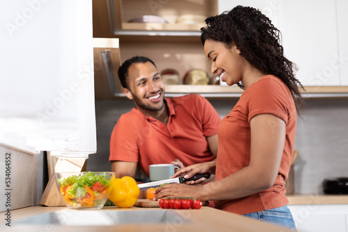 Happy millennial black man with coffee cup talking to his wife, lady cutting organic vegetables to salad in kitchen