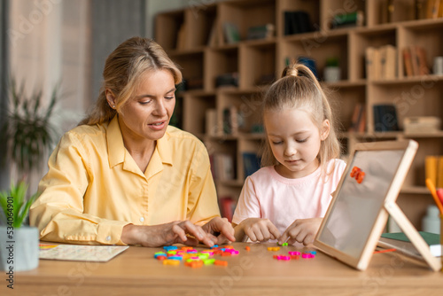 Friendly woman teaching pretty little girl alphabet, specialist working with child, making words from plastic letters