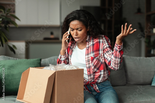Sad frustrated millennial african american woman opening cardboard box, talking on phone, swearing photo