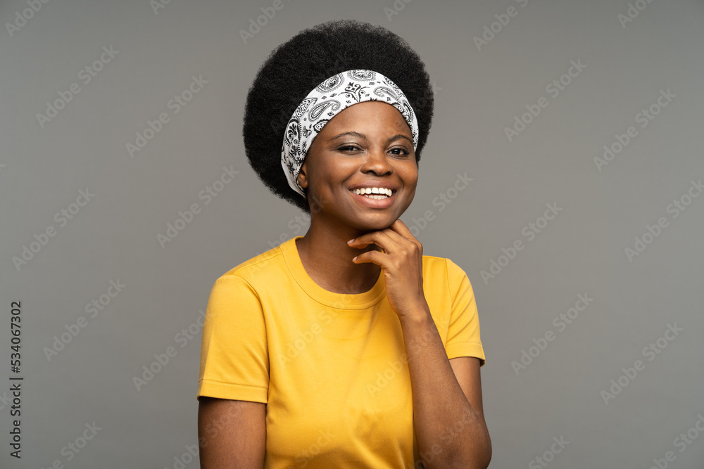 Happy African young woman wearing headband on kinky hair and yellow t-shirt demonstrates positive emotions performing at casting against grey studio wall. Black female enjoys life working cast member Happy African young woman wearing headband on kinky hair and yellow t-shirt demonstrates positive emotions performing at casting against grey studio wall. Black female enjoys life working cast member