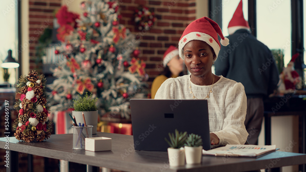 African american woman wearing santa hat and working in company office with christmas tree and festive lights. Female worker sending email on laptop during xmas winter holiday season. African american woman wearing santa hat and working in company office with christmas tree and festive lights. Female worker sending email on laptop during xmas winter holiday season.