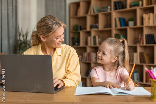 Happy mother and her little daughter spending time together, cute girl drawing while her mom working on laptop