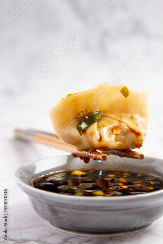 a gyosa on some Chinese chopsticks that is on a container of soy with seasoning on a white and gray marble background photo