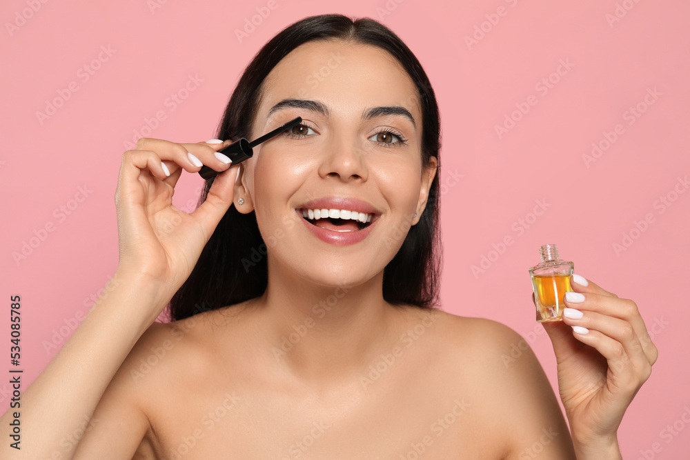 Fototapeta premium Young woman applying oil onto eyelashes on pink background