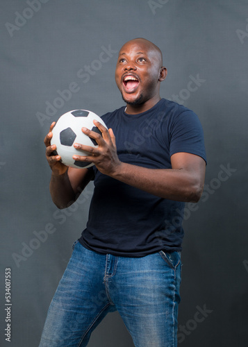 An excited African man or guy holding a black and white football with both his hands while excitedly celebrating with mouth oped wide and also looking up above