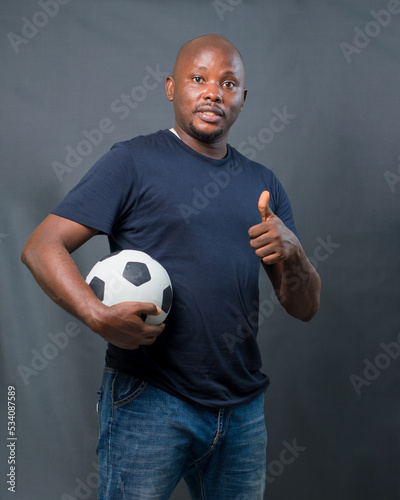 An excited African man or guy doing thumbs up gesture and holding a black and white football to his body while excitedly celebrating, jubilating and looking at the camera photo