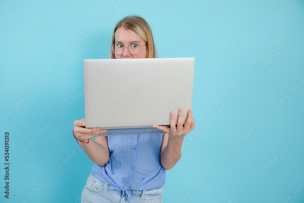 Portrait of shocked teenage girl with long hair wearing blue sleeveless shirt, holding laptop on blue background. Portrait of shocked teenage girl with long hair wearing blue sleeveless shirt, holding laptop on blue background.