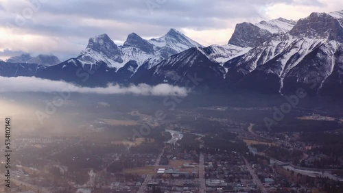 Slow dramatic left pan over town of Canmore in a valley in Alberta, Canada, as the sun rises over low clouds, with three sisters mountain range towering in the background. photo