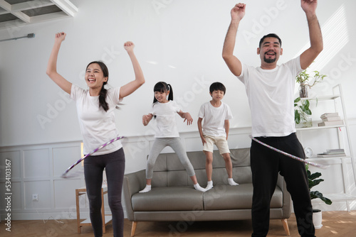 Happy wellness Asian Thai family, parents, and children are fun playing hula hoops together, fitness training and healthy exercise in white living room, domestic home lifestyle, and weekend activity.
