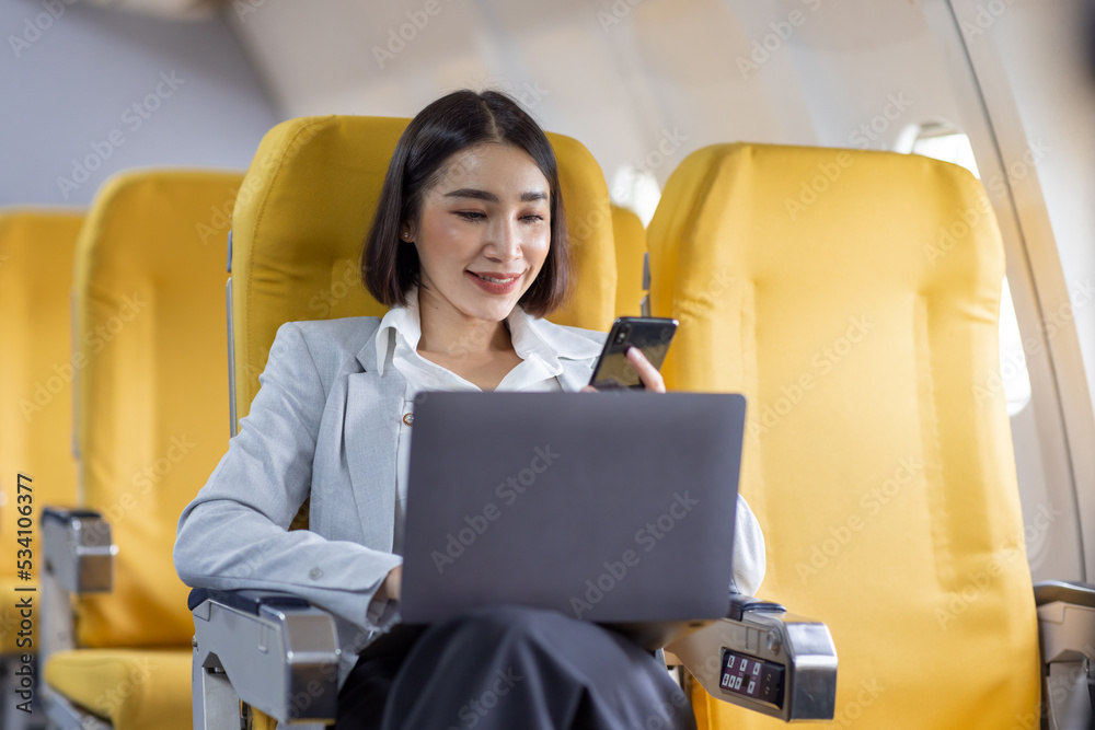 Asian female tourist checking incoming notification on smartphone and sitting on seat of airplane with laptop.Young Asian businesswoman share media from telephone on laptop during plane flight Asian female tourist checking incoming notification on smartphone and sitting on seat of airplane with laptop.Young Asian businesswoman share media from telephone on laptop during plane flight