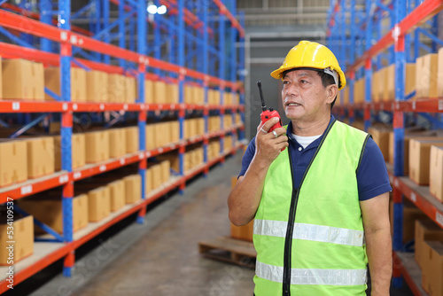 Asian warehouse worker man with hardhat and reflective jackets standing and using walkie talkie radio and cardboard while looking at something in retail warehouse logistics, distribution center