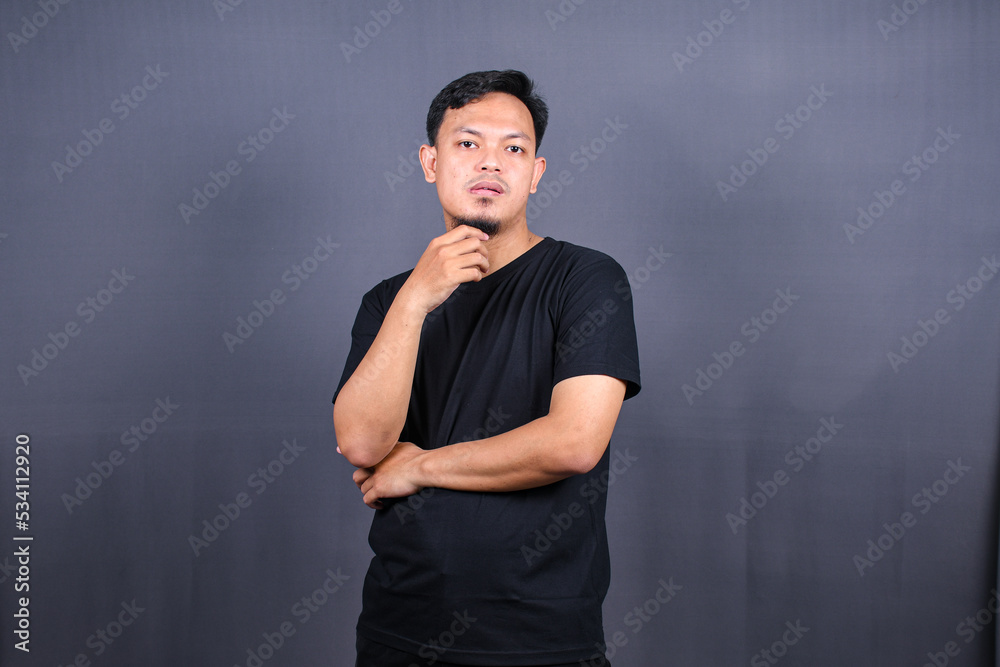 young Asian man holds chin and looks offended at camera feels angry awakes in bad mood isolated over gray background