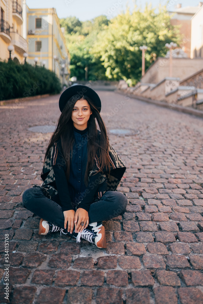 Naklejka premium Young woman in hat sitting at city on cobblestone