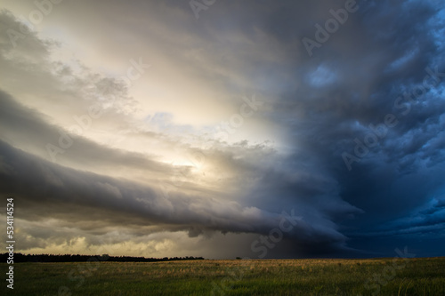 Sunlight partially illuminates an evening storm on the great plains with a tree line on the horizon.