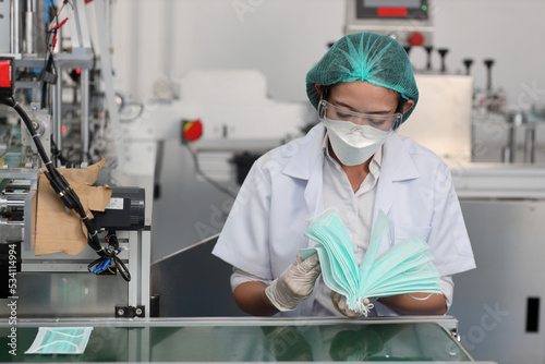 Worker woman in personal protective equipment or PPE inspecting quality of mask and medical face mask production line in factory  manufacturing industry and factory concept.