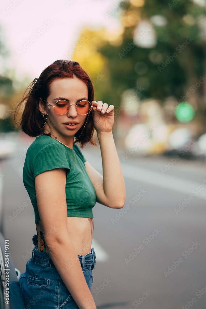 Beautiful fashionable young woman in glasses posing near car