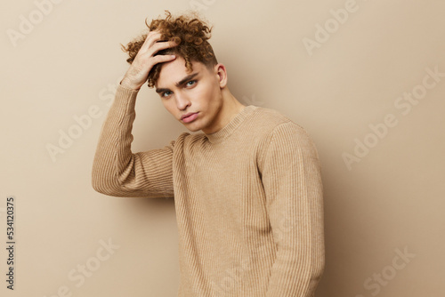 a charming, handsome man stands on a beige background in a sweatshirt and looks relaxed, holding his hands behind his head with his elbows apart