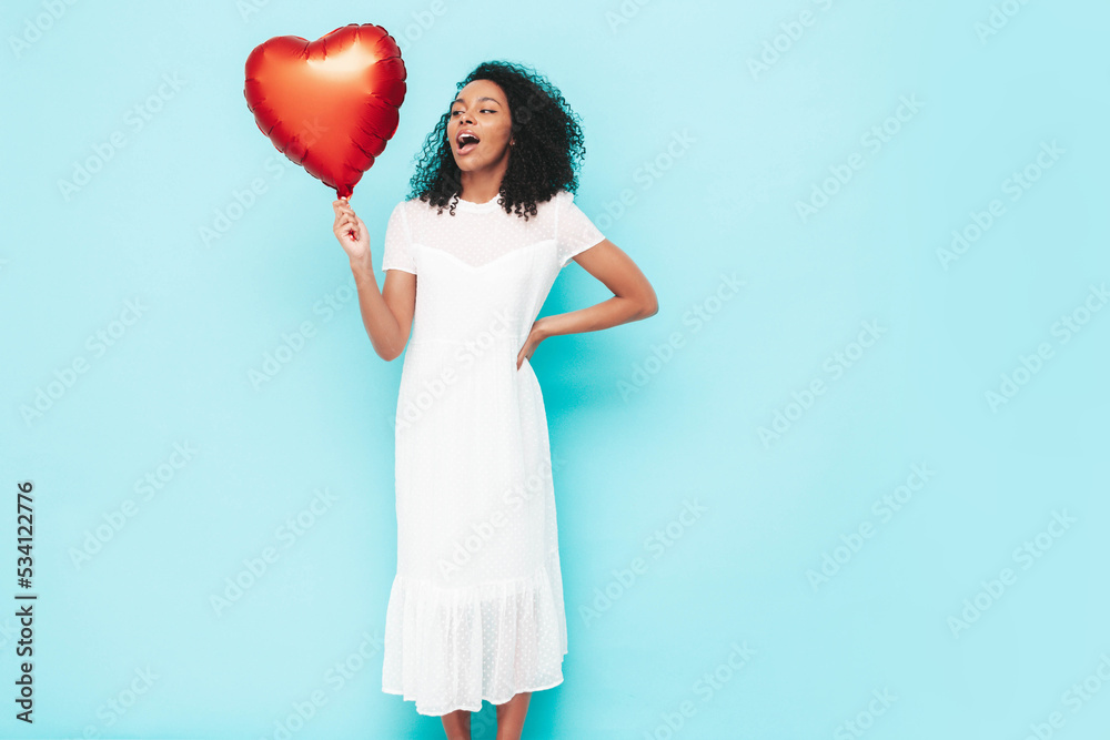 Beautiful black woman with afro curls hairstyle. Smiling model dressed in white summer dress. Sexy carefree female posing near blue wall in studio. Tanned and cheerful. Holding heart air balloon Beautiful black woman with afro curls hairstyle. Smiling model dressed in white summer dress. Sexy carefree female posing near blue wall in studio. Tanned and cheerful. Holding heart air balloon