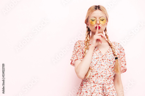 Young beautiful smiling female in trendy summer dress. Sexy carefree blond  woman posing near wall in studio. Cheerful and happy in sunglasses. Shows hush sign, silence