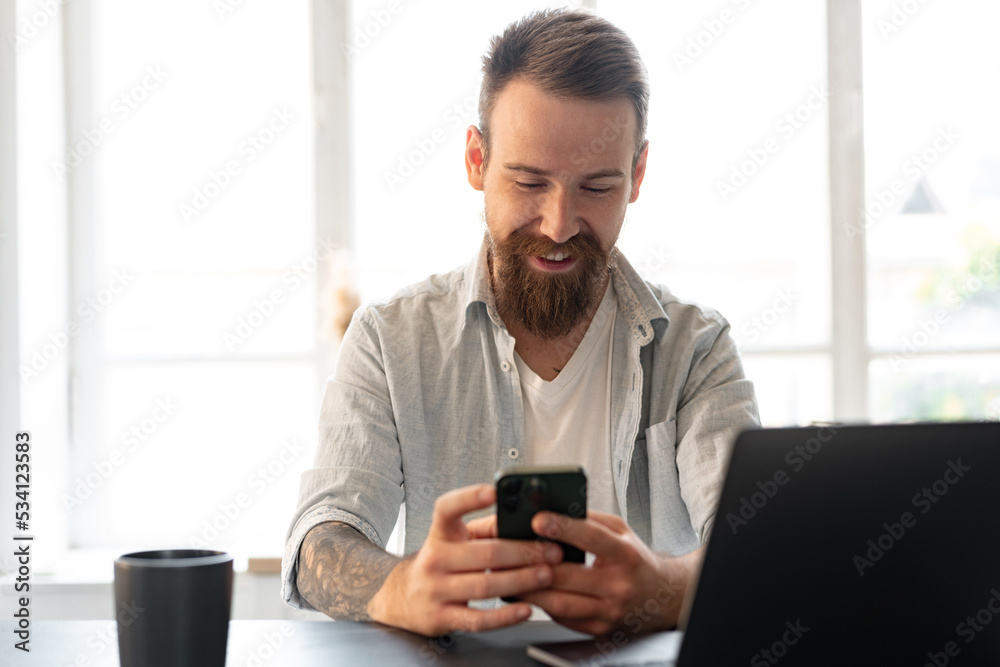 Handsome young bearded man spending time at home with phone in hands.