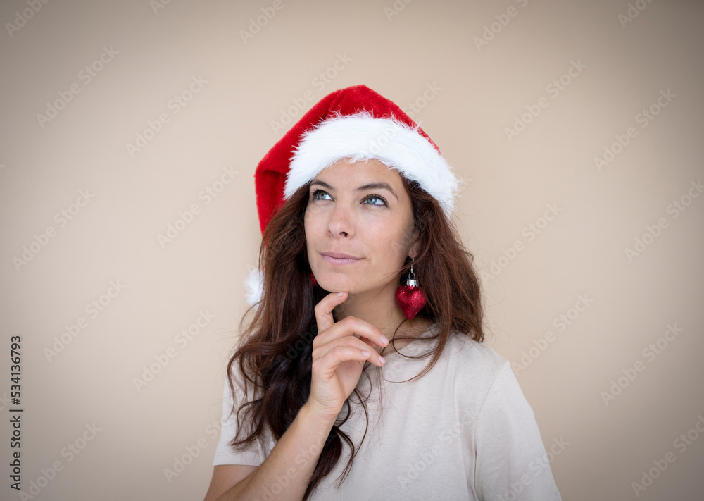 Pretty young woman with dark hair wearing Christmas hat and heart earrings in the shape of Christmas tree ball in front of brown background Pretty young woman with dark hair wearing Christmas hat and heart earrings in the shape of Christmas tree ball in front of brown background
