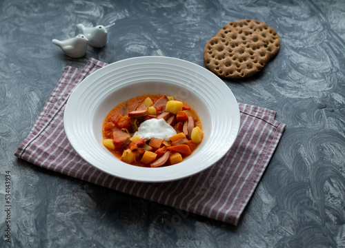 Fototapeta Naklejka Na Ścianę i Meble -  Soup with sausages and sour cream in a white plate on a striped napkin. Two slices of bread.