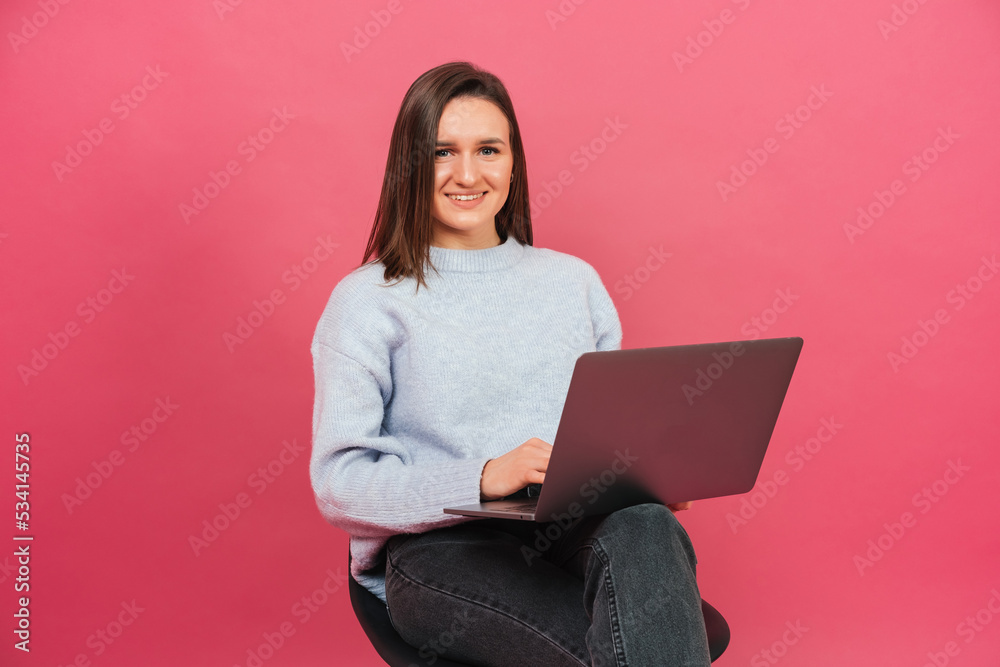 Naklejka premium Photo of young woman sitting on chair and using laptop over pink background.