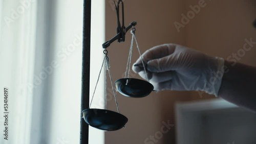 Close-up of medical worker hand in rubber gloves measuring medicine, putting weight on traditional metal balance scale, scales moving due imbalance. Concept of weights comparison.