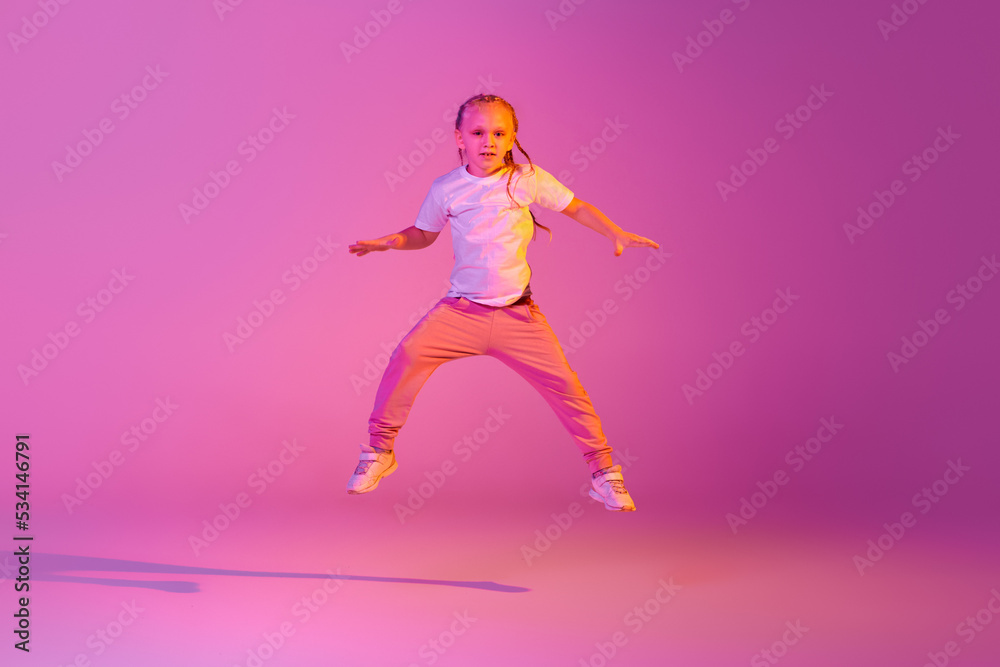 Beautiful active little girl, kid jumping, dancing isolated over pink background in neon. Action, dance, happy childhood