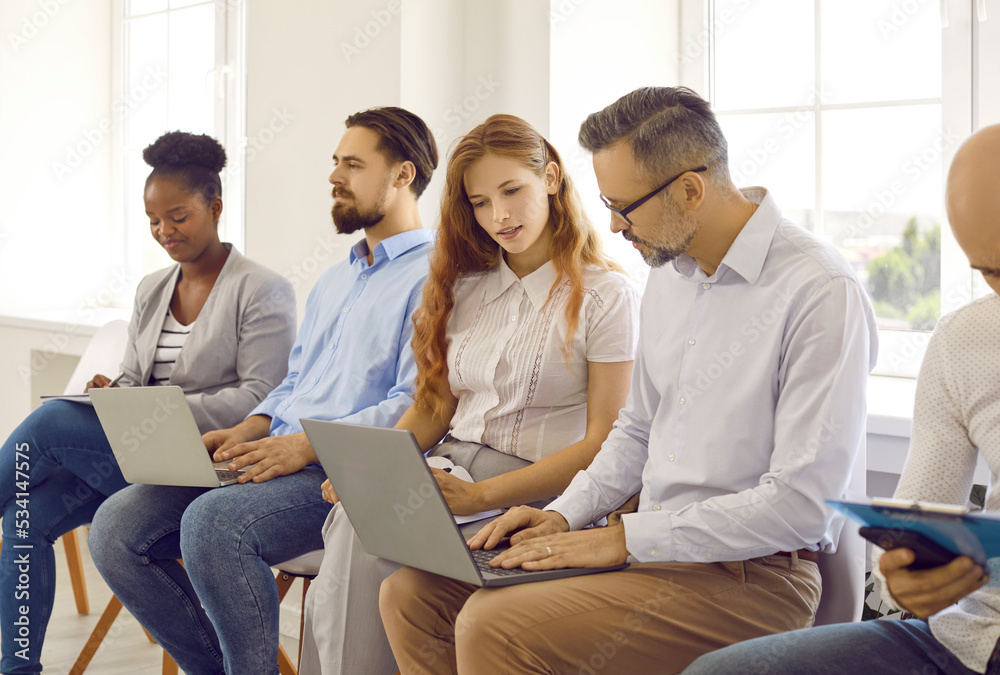 Diverse people using laptop computers while sitting in line in office workplace. Group of multiracial young and mature men and women sitting in row, using notebook PCs and working on business projects Diverse people using laptop computers while sitting in line in office workplace. Group of multiracial young and mature men and women sitting in row, using notebook PCs and working on business projects