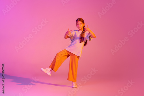 Moves. Cute little girl, kid in casual bright clothes dancing isolated over pink background in neon. Action, dance, happy childhood