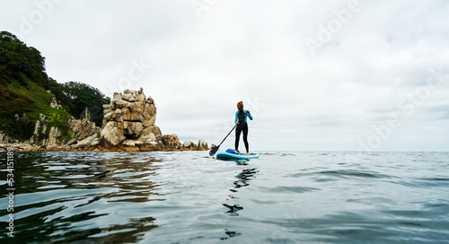 Woman in wetsuit stand up on paddle board in sea. Big blue board in turquoise water.