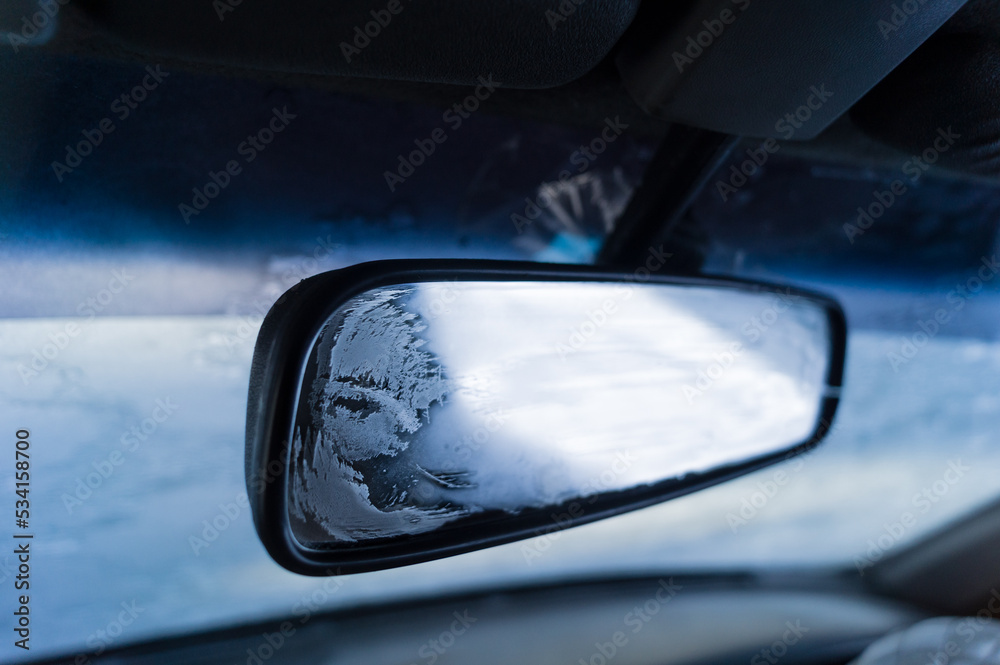An icy rear-view mirror inside the cabin of a frozen car. Problem of icing of the windows and mirrors of the car. Ice on the rear-view mirror. Low air temperature inside the cabin. Selective focus